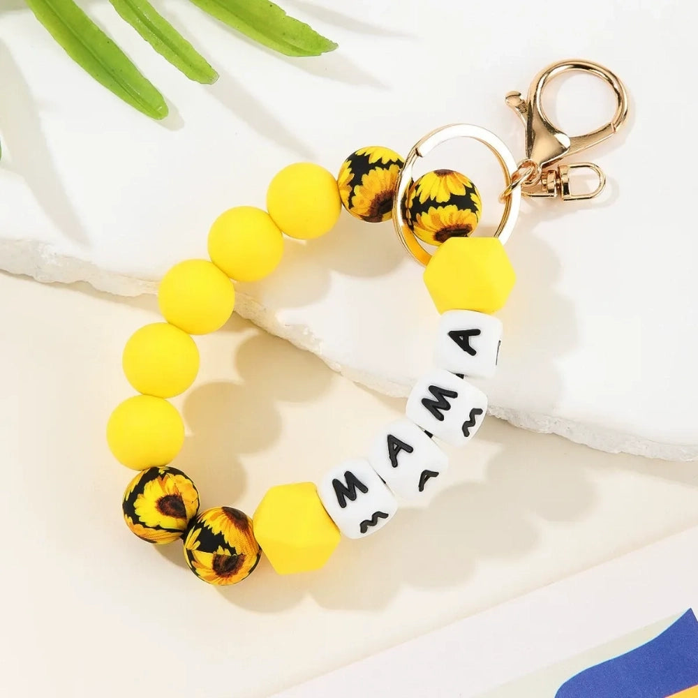 Seven MAMA letter silicone bead wristlet keychains in assorted colors including mint, lavender, gray, yellow, pink, black, and green with patterned accent beads and gold lobster clasp hardware on a light background. - Yellow