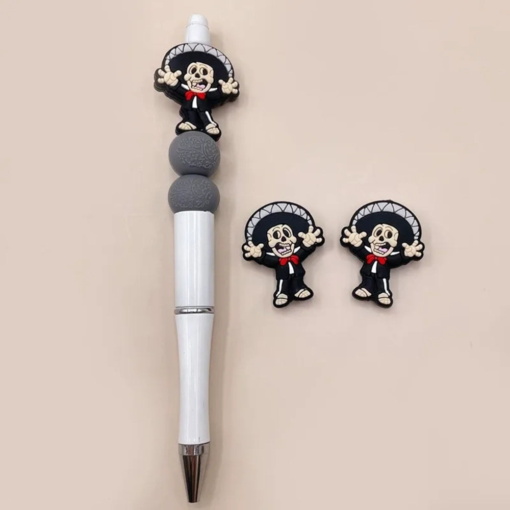 Mariachi skeleton silicone focal bead featuring a Day of the Dead skull in a black sombrero suit with red bow tie, shown on a beadable pen with two loose flatback focal charms on a beige background.