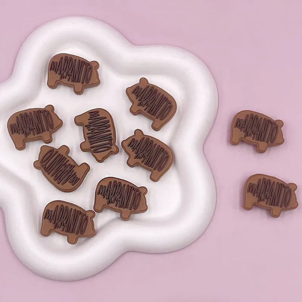 Brown silicone focal bead shaped like a pig with 'Marranito' text embossed on body, shown in multiple pieces on a white wavy tray with pink background.