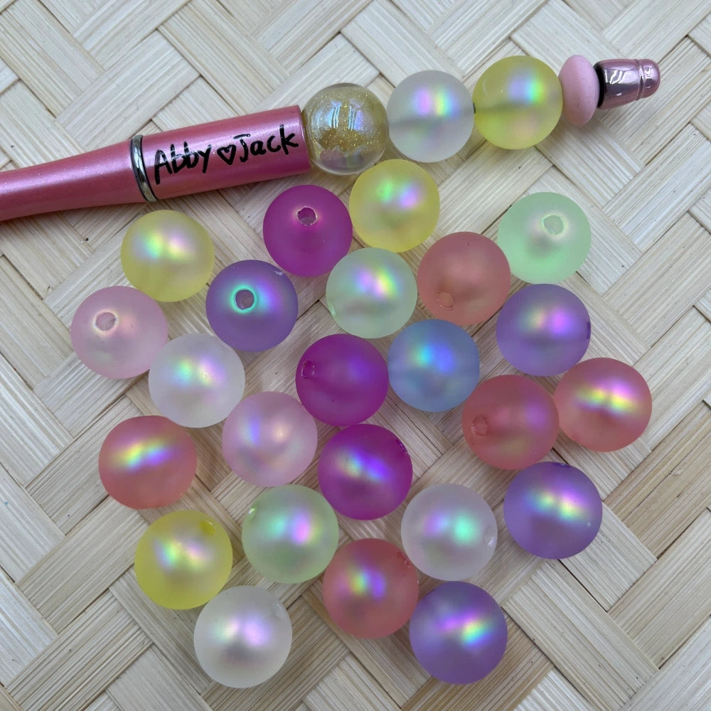 Matte aurora iridescent round acrylic beads in pastel pink, purple, yellow, and white with rainbow shimmer, displayed next to a beadable pen on a woven surface.