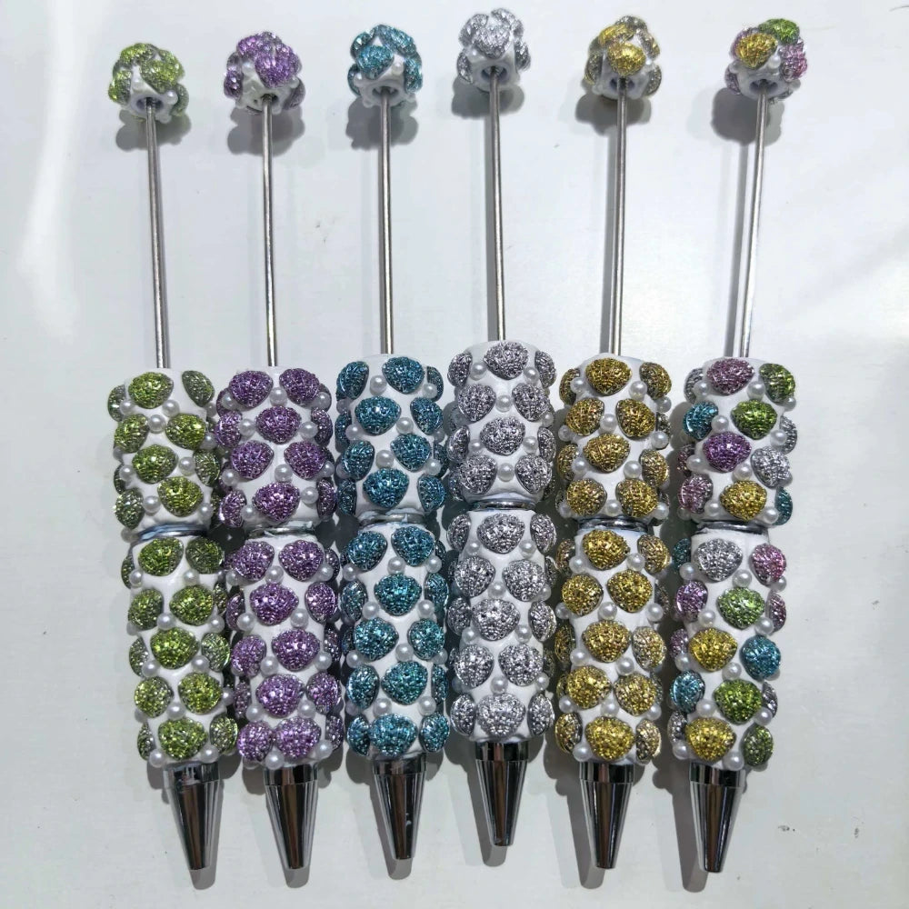 Six decorative beadable pens featuring glitter rhinestone clay bumps and pearl accents in assorted colors including green, purple, teal, silver, gold, and mixed on white barrel with silver metal tip and topper bead.