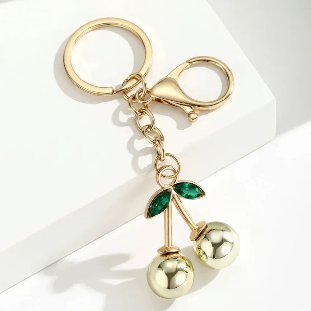 Six colorful metallic cherry bell charm keychains in pink, silver, blue, gold, red, and light pink with green crystal leaves and gold lobster clasp hardware on white background. - 1