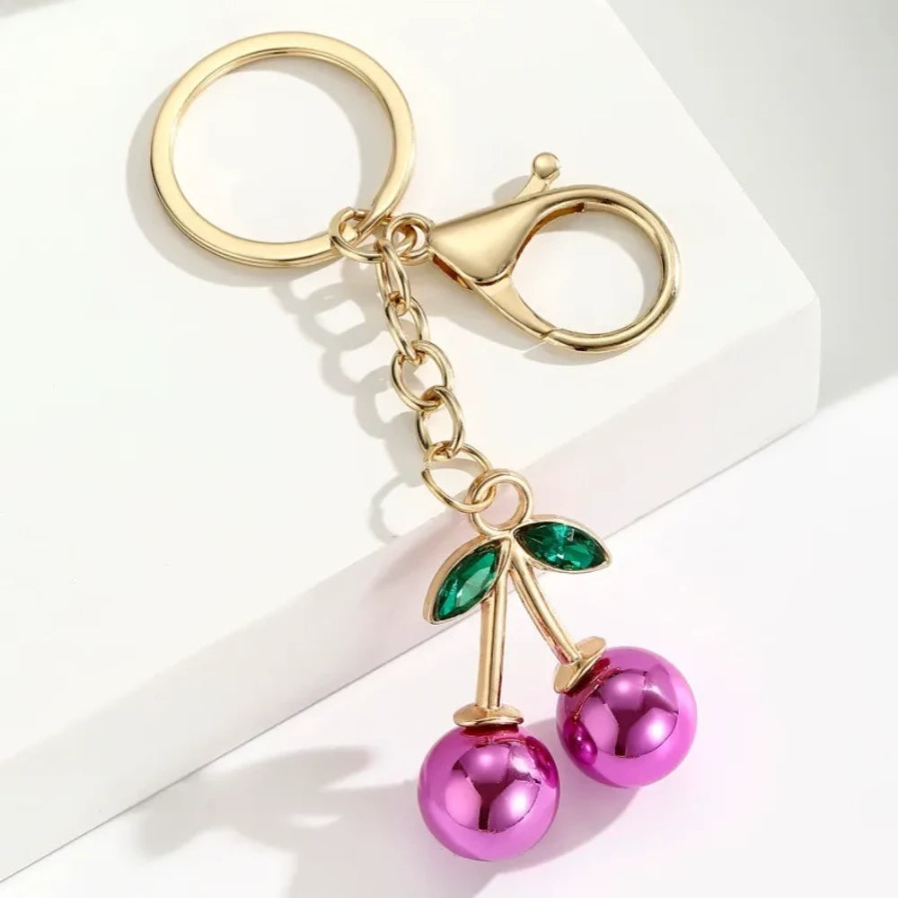 Six colorful metallic cherry bell charm keychains in pink, silver, blue, gold, red, and light pink with green crystal leaves and gold lobster clasp hardware on white background. - 2