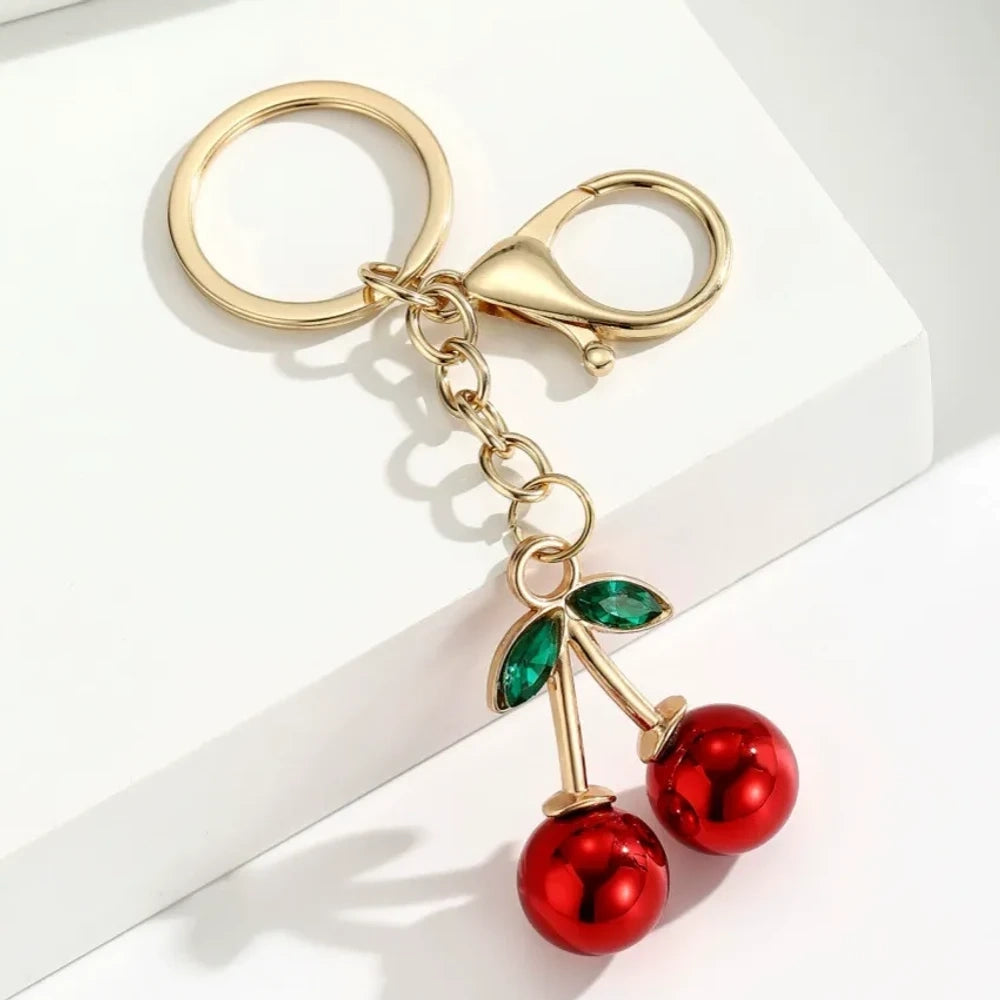Six colorful metallic cherry bell charm keychains in pink, silver, blue, gold, red, and light pink with green crystal leaves and gold lobster clasp hardware on white background. - 3