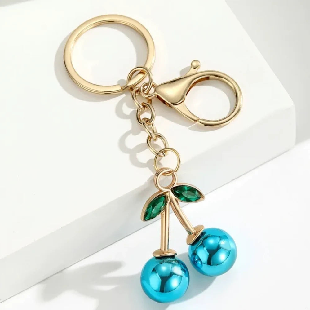 Six colorful metallic cherry bell charm keychains in pink, silver, blue, gold, red, and light pink with green crystal leaves and gold lobster clasp hardware on white background. - 4