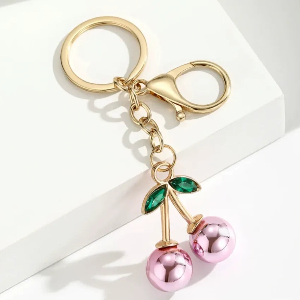 Six colorful metallic cherry bell charm keychains in pink, silver, blue, gold, red, and light pink with green crystal leaves and gold lobster clasp hardware on white background. - 5