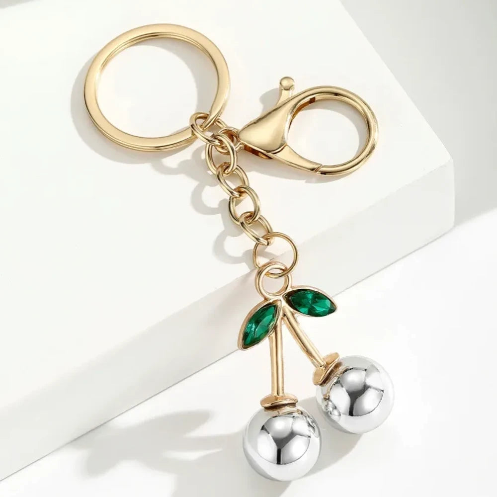Six colorful metallic cherry bell charm keychains in pink, silver, blue, gold, red, and light pink with green crystal leaves and gold lobster clasp hardware on white background. - 6