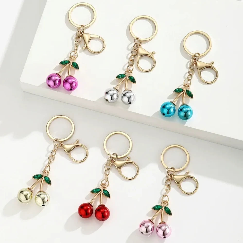 Six colorful metallic cherry bell charm keychains in pink, silver, blue, gold, red, and light pink with green crystal leaves and gold lobster clasp hardware on white background.