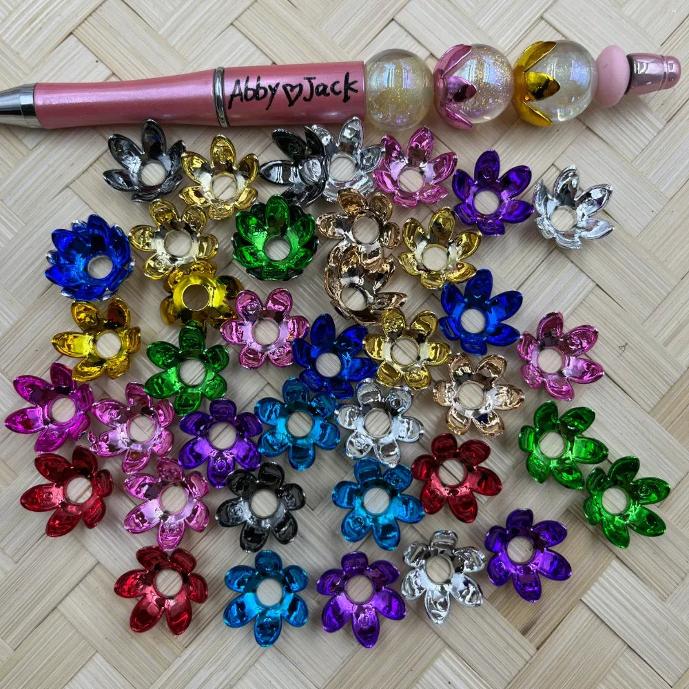 Assorted metallic flower-shaped bead caps in multiple colors including blue, gold, green, purple, pink, red, silver, and black, displayed on a woven surface next to a beadable pen labeled Abby and Jack.