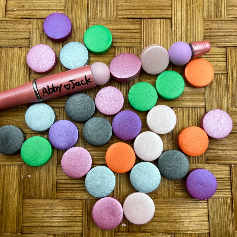 Colorful metallic shimmer flat round coin beads in purple, green, orange, pink, teal, white, and gray scattered on a wicker surface next to a beadable pen labeled Abby and Jack.