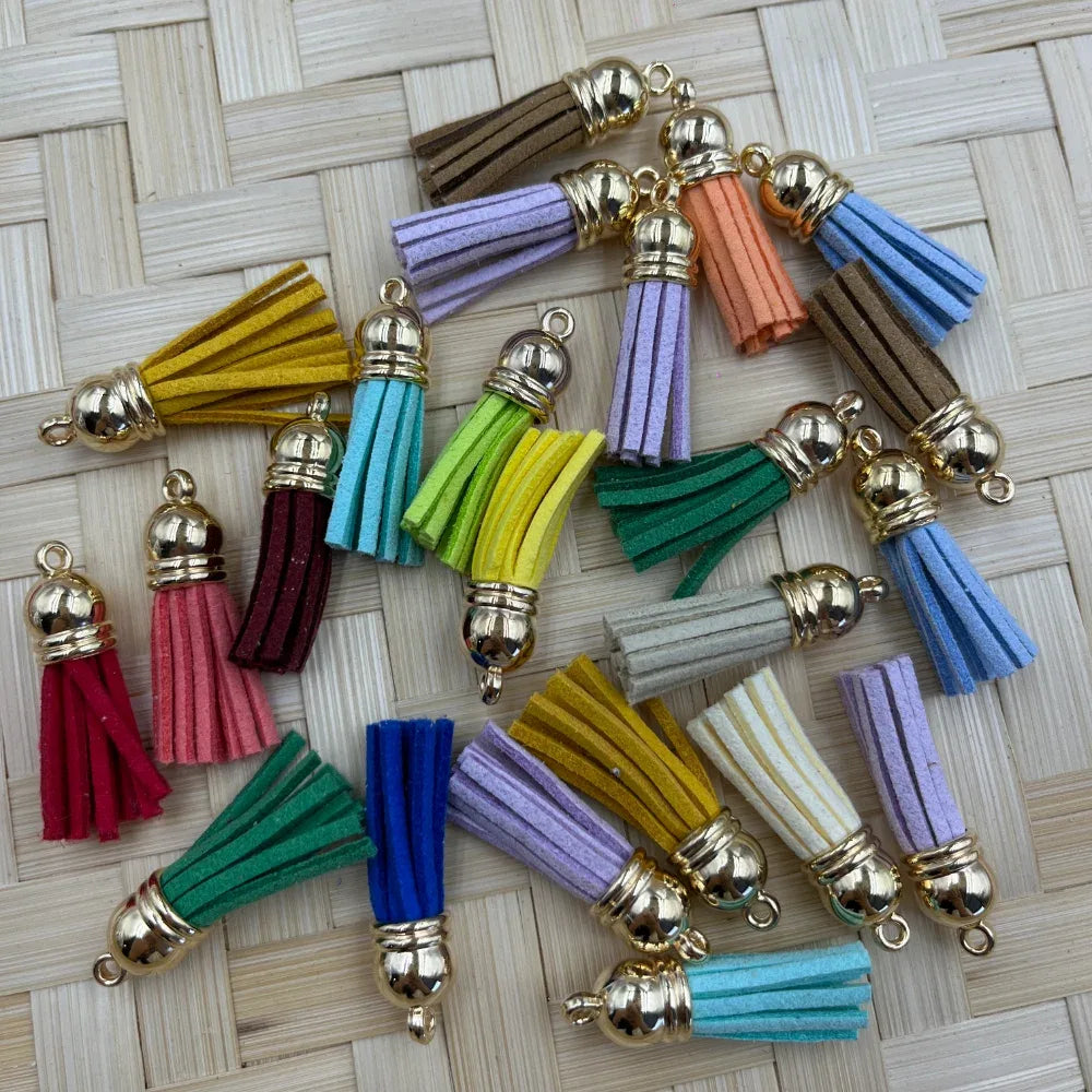 Assorted colorful mini suede tassel charms with gold metal caps displayed on woven rattan surface, available in rainbow of colors including pink, purple, green, blue, yellow and more.