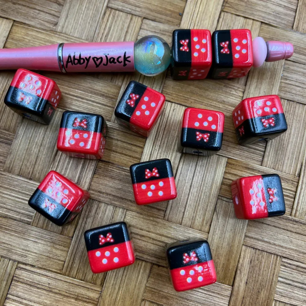 Red and black Minnie Mouse bow polka dot acrylic cube beads displayed with a beadable pen on a wicker surface, showing white dot pip details and red bow accents on each bead face.