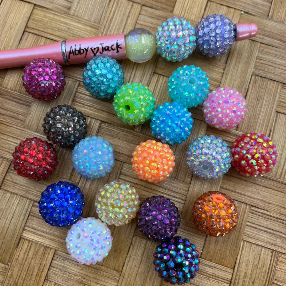 Assorted mixed color AB rhinestone sparkle beads in pink, teal, blue, purple, orange, red, green, white, yellow and more, displayed on a woven surface next to a beadable pen.