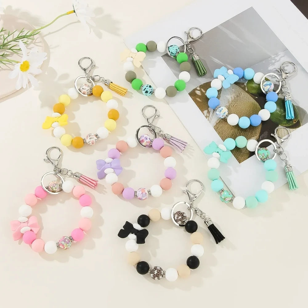 Set of colorful silicone bead stretch bracelet keychains in pastel and bold colors including pink, blue, green, yellow, purple, teal, and black, each featuring a lobster clasp, decorative printed silicone bead, tassel or bow accent, displayed on a white surface.