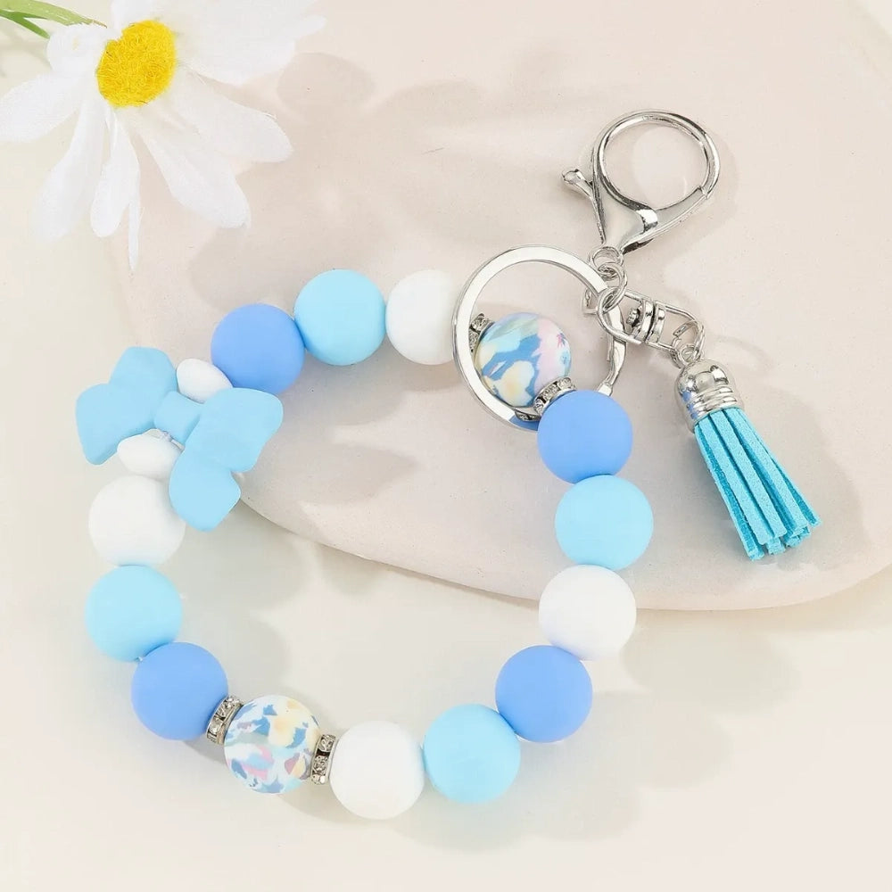 Set of colorful silicone bead stretch bracelet keychains in pastel and bold colors including pink, blue, green, yellow, purple, teal, and black, each featuring a lobster clasp, decorative printed silicone bead, tassel or bow accent, displayed on a white surface. - Blue