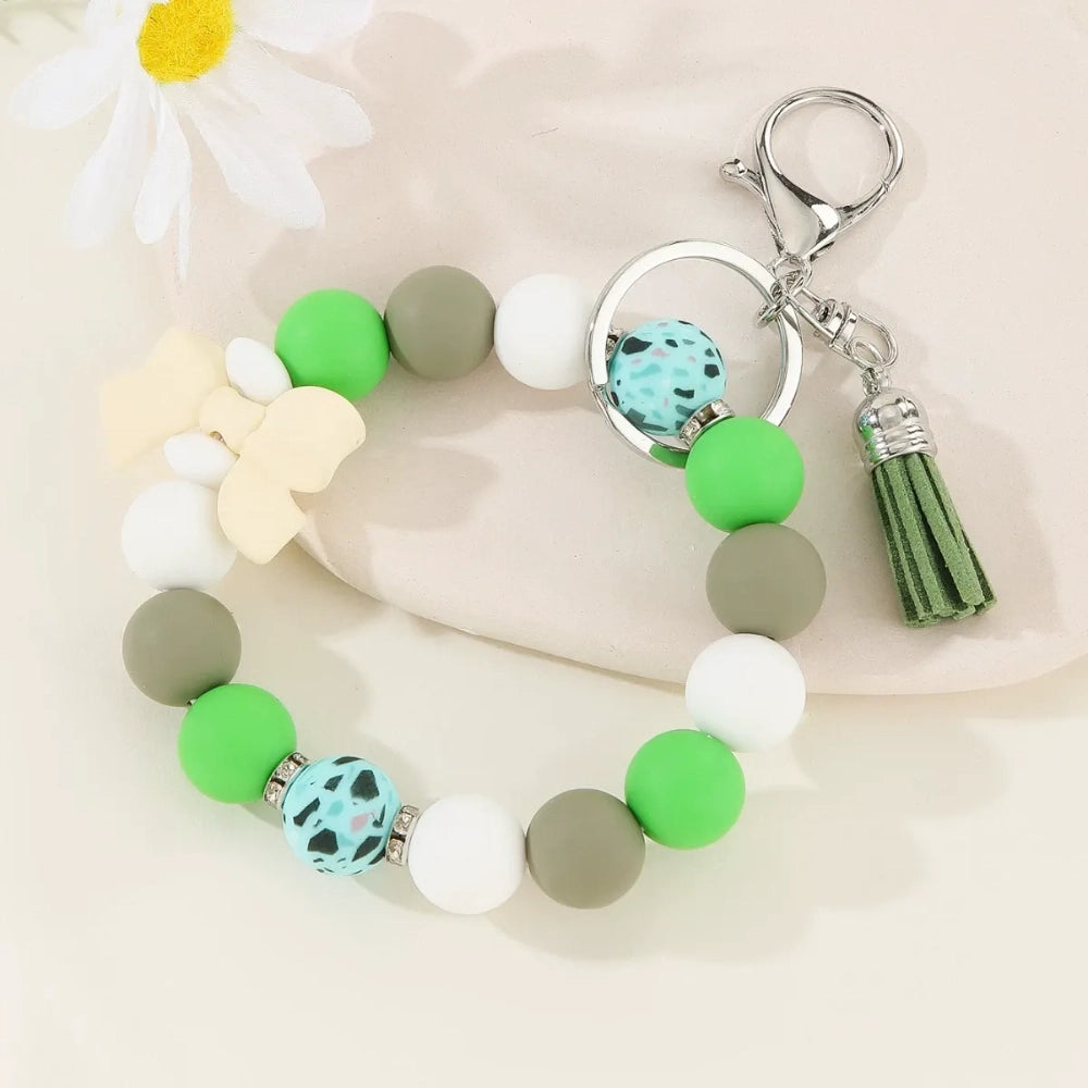 Set of colorful silicone bead stretch bracelet keychains in pastel and bold colors including pink, blue, green, yellow, purple, teal, and black, each featuring a lobster clasp, decorative printed silicone bead, tassel or bow accent, displayed on a white surface. - Green