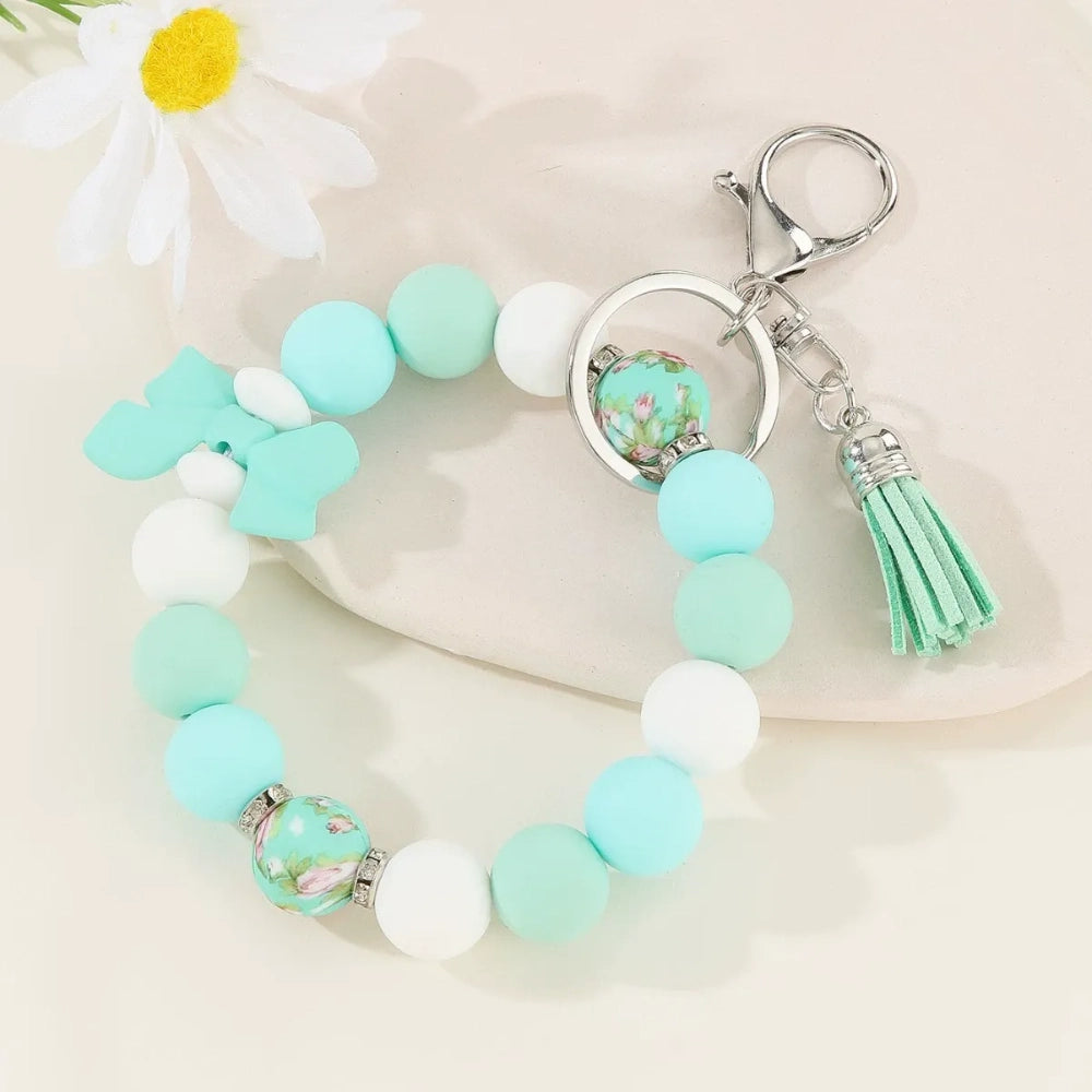 Set of colorful silicone bead stretch bracelet keychains in pastel and bold colors including pink, blue, green, yellow, purple, teal, and black, each featuring a lobster clasp, decorative printed silicone bead, tassel or bow accent, displayed on a white surface. - Light Green