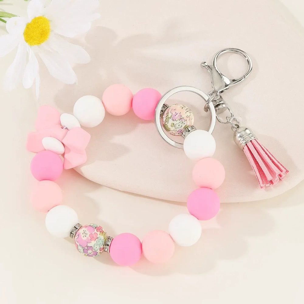 Set of colorful silicone bead stretch bracelet keychains in pastel and bold colors including pink, blue, green, yellow, purple, teal, and black, each featuring a lobster clasp, decorative printed silicone bead, tassel or bow accent, displayed on a white surface. - Pink