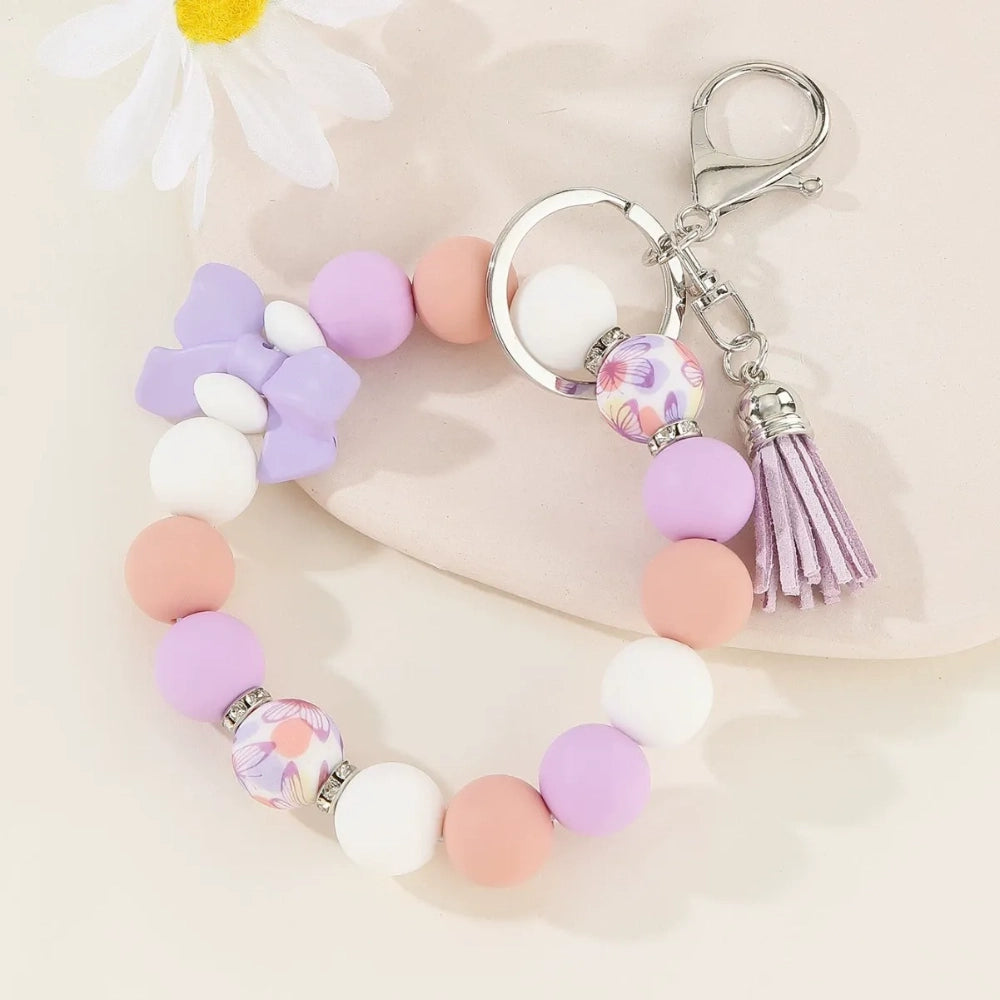 Set of colorful silicone bead stretch bracelet keychains in pastel and bold colors including pink, blue, green, yellow, purple, teal, and black, each featuring a lobster clasp, decorative printed silicone bead, tassel or bow accent, displayed on a white surface. - Purple