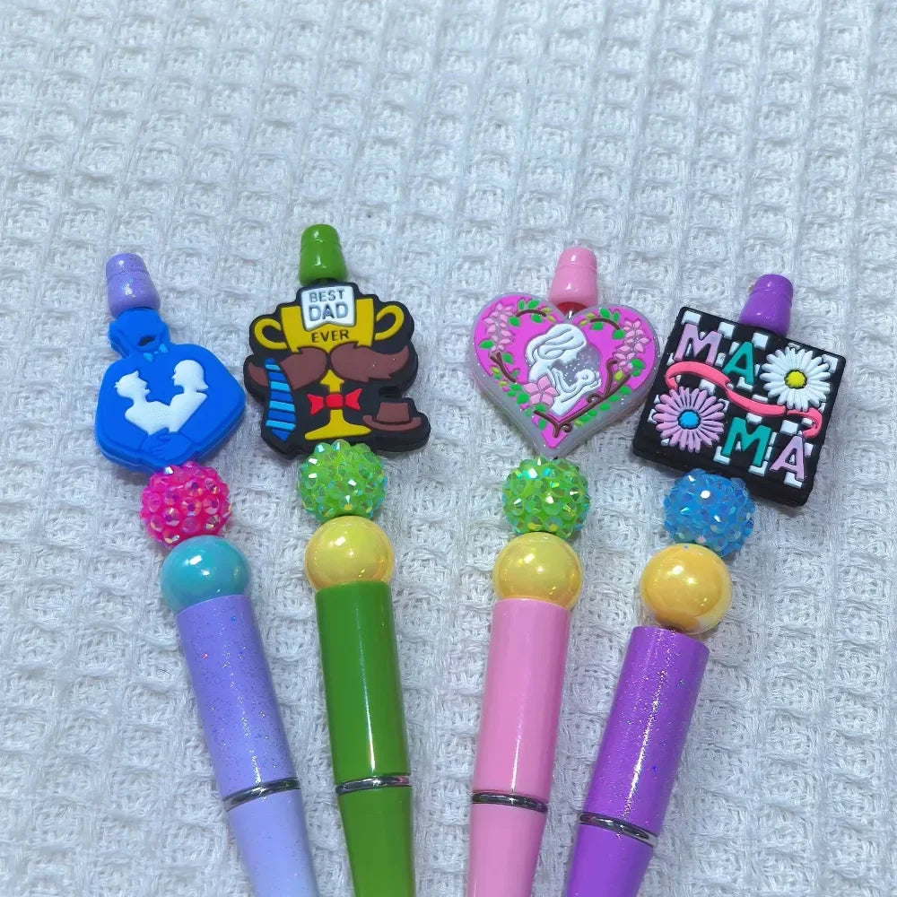 Four silicone focal beads featuring Mom and Dad themed designs including Best Dad Ever trophy, Mama text, mother and child heart, and father silhouette, displayed on decorated beadable pens in purple, green, pink, and purple colors.