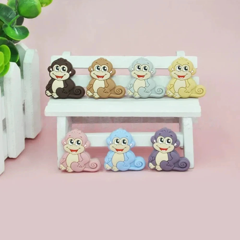Seven colorful cartoon monkey silicone focal beads in brown, tan, grey, yellow, pink, blue, and purple displayed on a miniature white bench against a pink background.