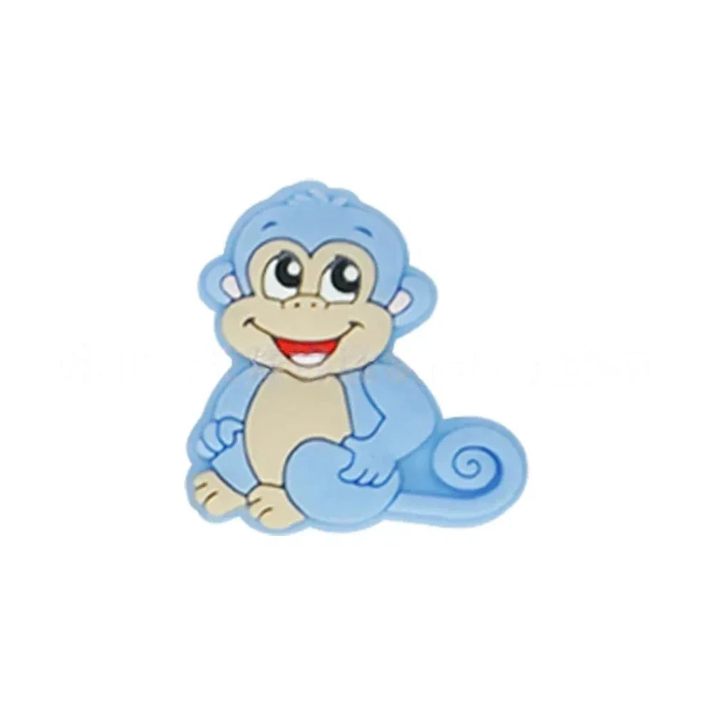 Seven colorful cartoon monkey silicone focal beads in brown, tan, grey, yellow, pink, blue, and purple displayed on a miniature white bench against a pink background. - Blue