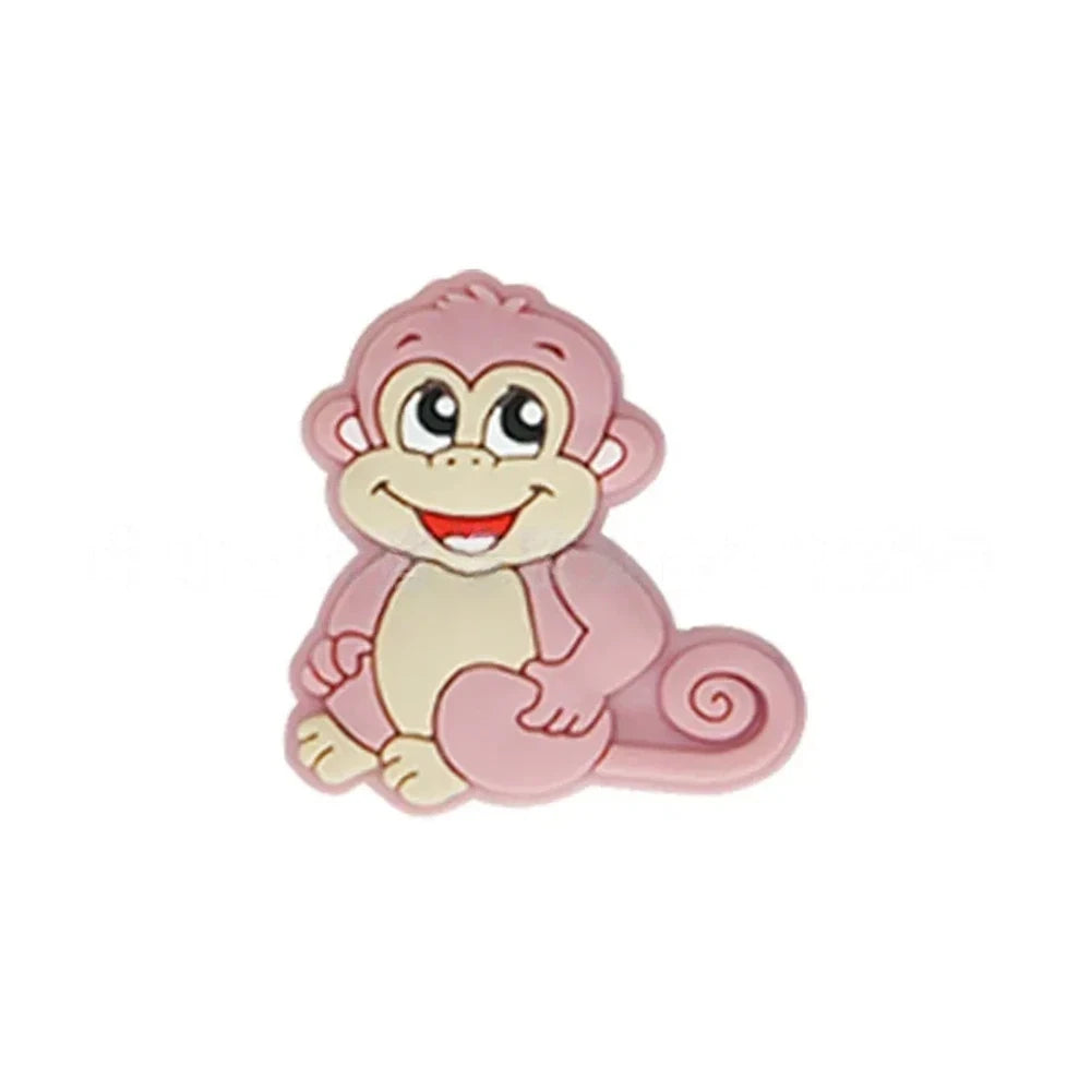 Seven colorful cartoon monkey silicone focal beads in brown, tan, grey, yellow, pink, blue, and purple displayed on a miniature white bench against a pink background. - Pink