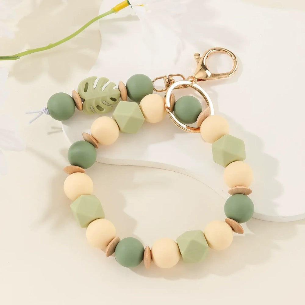 Monstera leaf silicone bead bracelet keychains in sage green with cream beads and teal blue with mint beads, featuring gold lobster clasp hardware on a white surface with tropical leaves. - 1