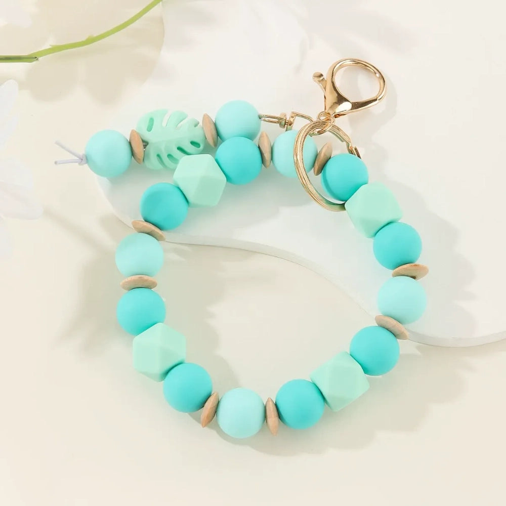 Monstera leaf silicone bead bracelet keychains in sage green with cream beads and teal blue with mint beads, featuring gold lobster clasp hardware on a white surface with tropical leaves. - 2