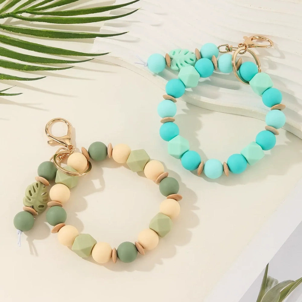 Monstera leaf silicone bead bracelet keychains in sage green with cream beads and teal blue with mint beads, featuring gold lobster clasp hardware on a white surface with tropical leaves. - Random Mixed