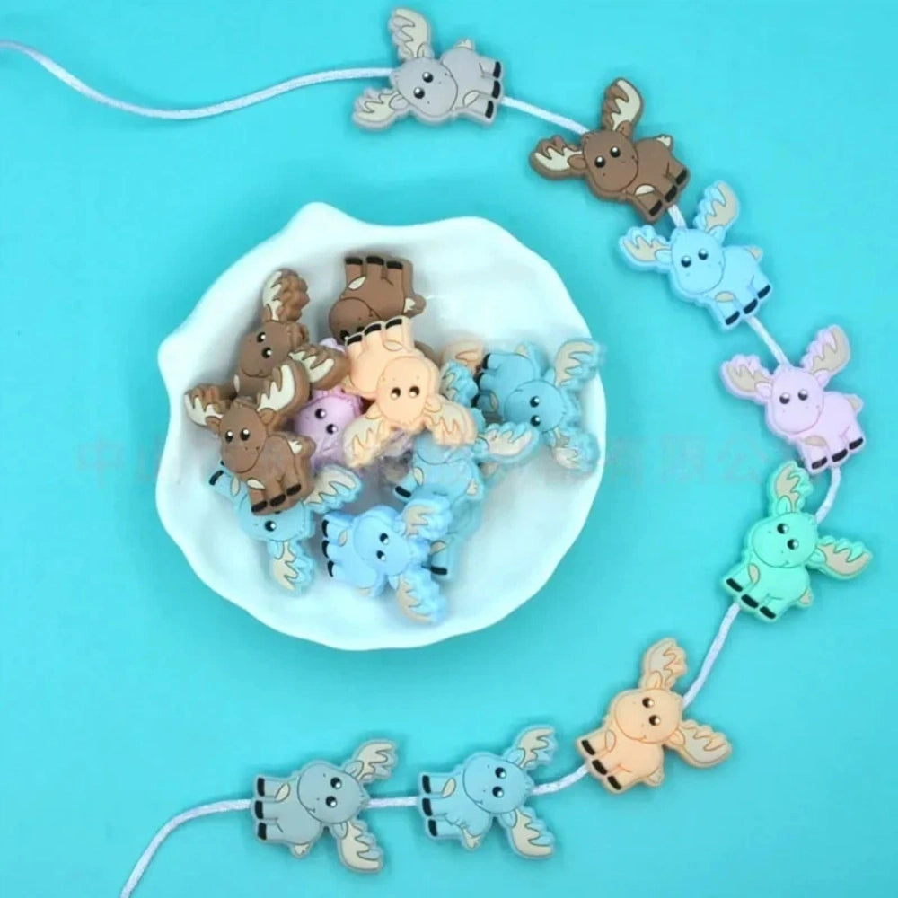 Assorted pastel and earth tone moose silicone focal beads strung on a cord and displayed in a white dish on a teal background, ideal for beadable pens, keychains, and badge reels.