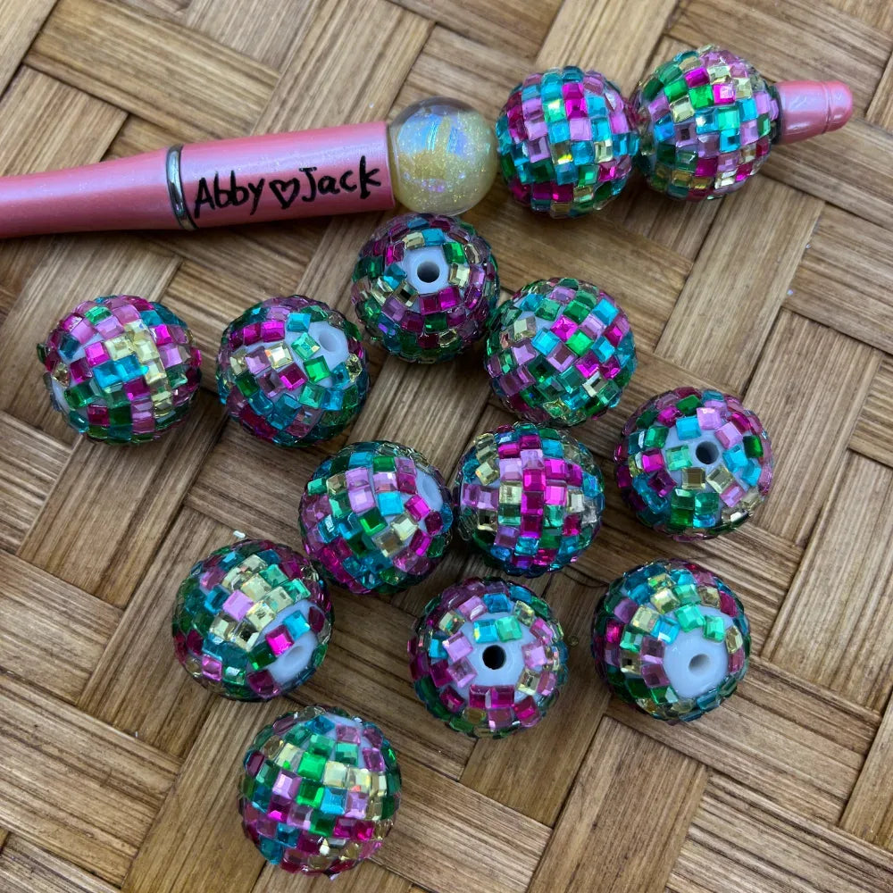 Multicolor disco ball rhinestone beads covered in pink, teal, green, and gold square rhinestones, displayed on a woven surface next to a beadable pen.