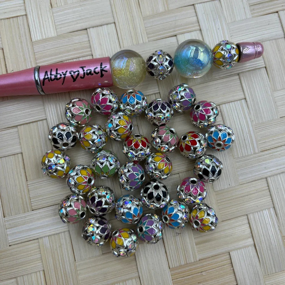 Multicolor enamel and AB rhinestone silver filigree round metal spacer beads with floral cutout design, displayed on woven bamboo mat next to a pink beadable pen labeled Abby Jack.