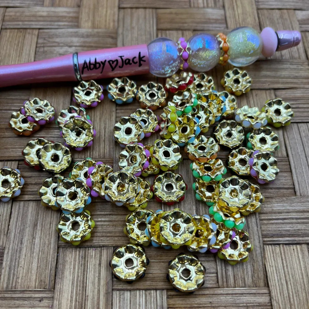 Gold rondelle metal spacer beads with multicolor rhinestone crystal edges scattered on a wicker surface next to a decorated beadable pen labeled Abby and Jack.