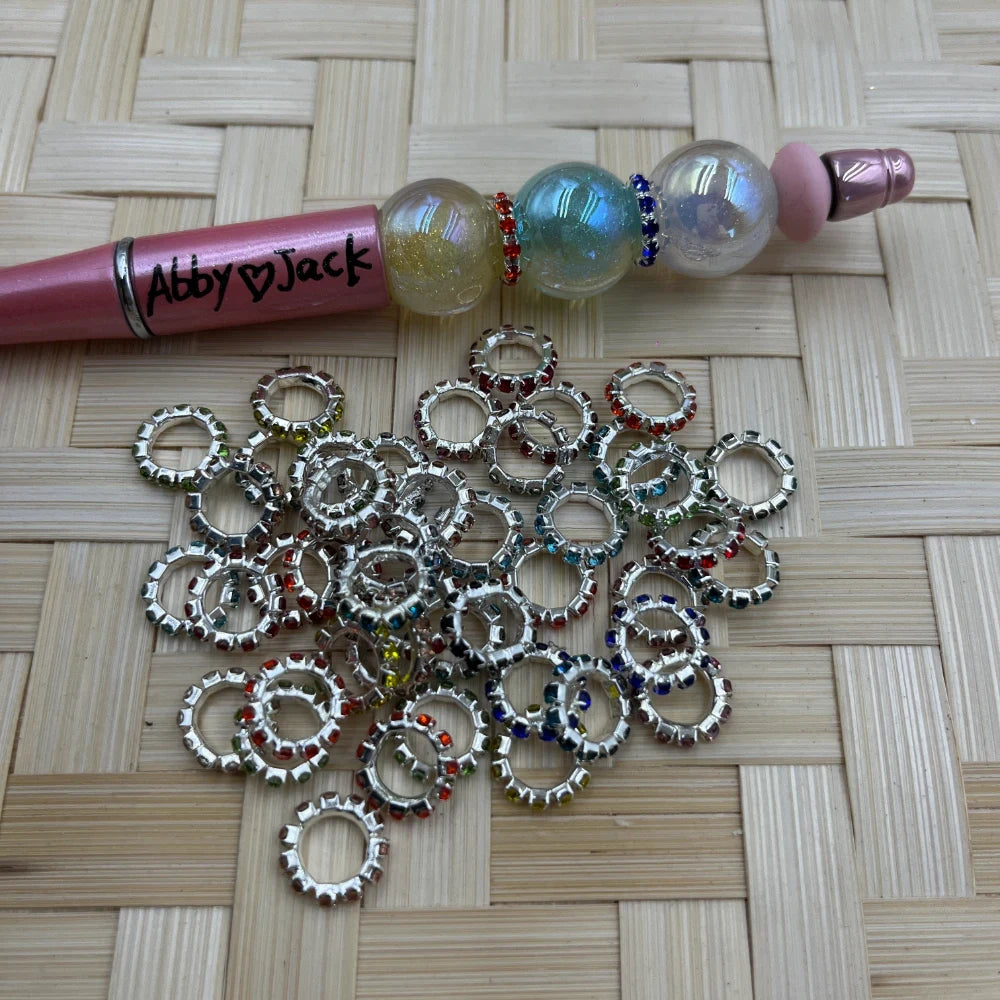 Assorted multicolor rhinestone ring metal spacer beads in silver tone scattered on woven surface next to a pink beadable pen labeled Abby Jack with iridescent globe beads.