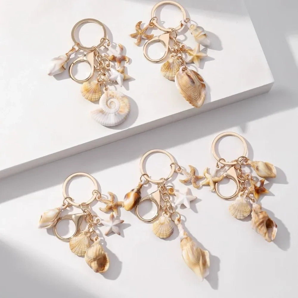 Five natural seashell keychain charms featuring conch shells, starfish, and scallop shells on gold-tone key rings, arranged on a white surface for DIY jewelry making and beach-themed crafts.