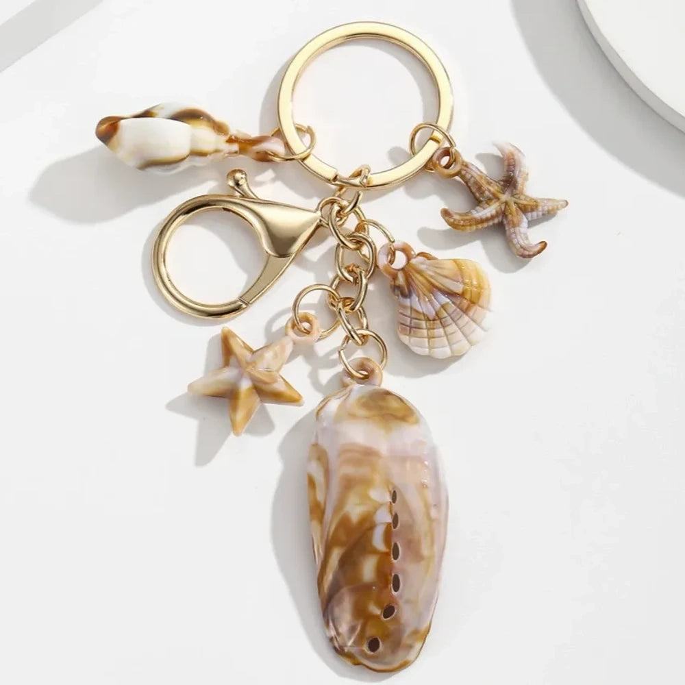 Five natural seashell keychain charms featuring conch shells, starfish, and scallop shells on gold-tone key rings, arranged on a white surface for DIY jewelry making and beach-themed crafts. - 1