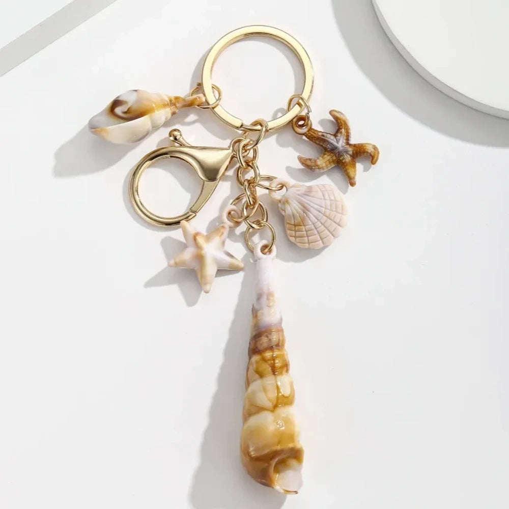 Five natural seashell keychain charms featuring conch shells, starfish, and scallop shells on gold-tone key rings, arranged on a white surface for DIY jewelry making and beach-themed crafts. - 2