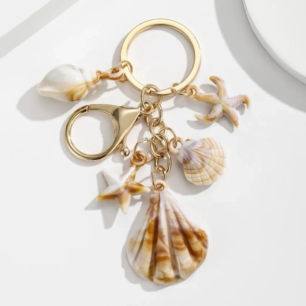 Five natural seashell keychain charms featuring conch shells, starfish, and scallop shells on gold-tone key rings, arranged on a white surface for DIY jewelry making and beach-themed crafts. - 5
