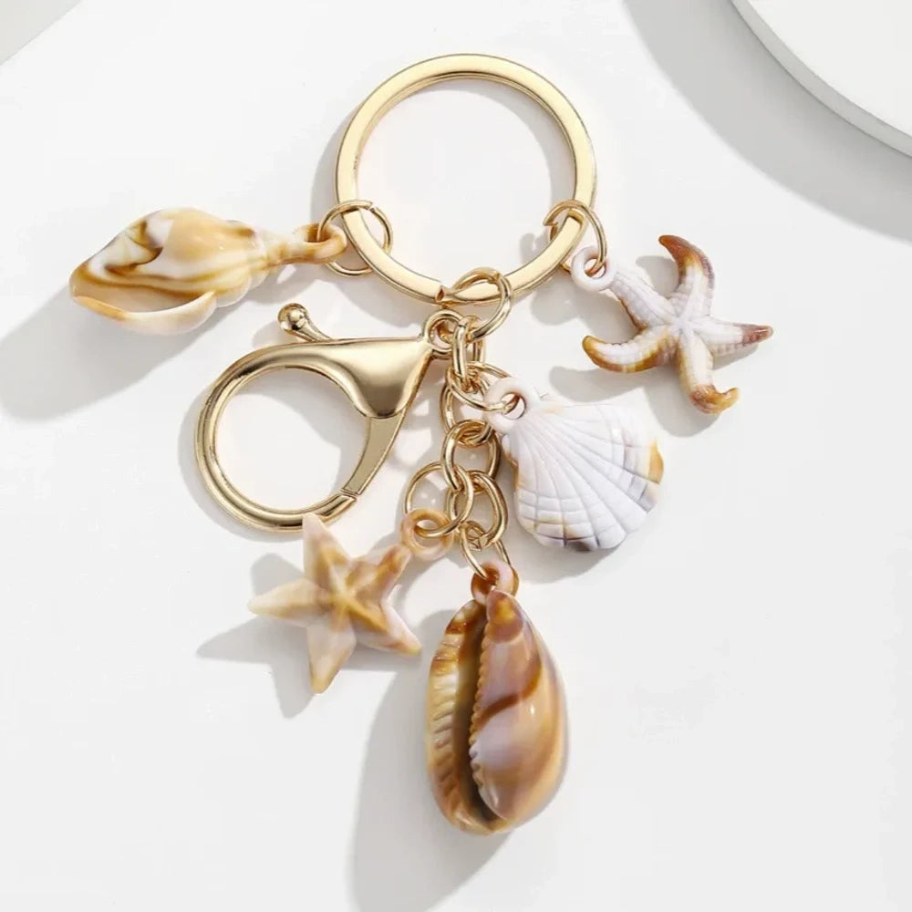 Five natural seashell keychain charms featuring conch shells, starfish, and scallop shells on gold-tone key rings, arranged on a white surface for DIY jewelry making and beach-themed crafts. - 6