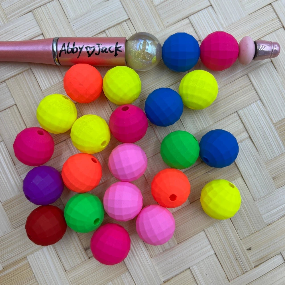 Assorted neon faceted acrylic beads in bright colors including yellow, pink, orange, blue, green, and purple displayed next to a beadable pen on a wicker surface.