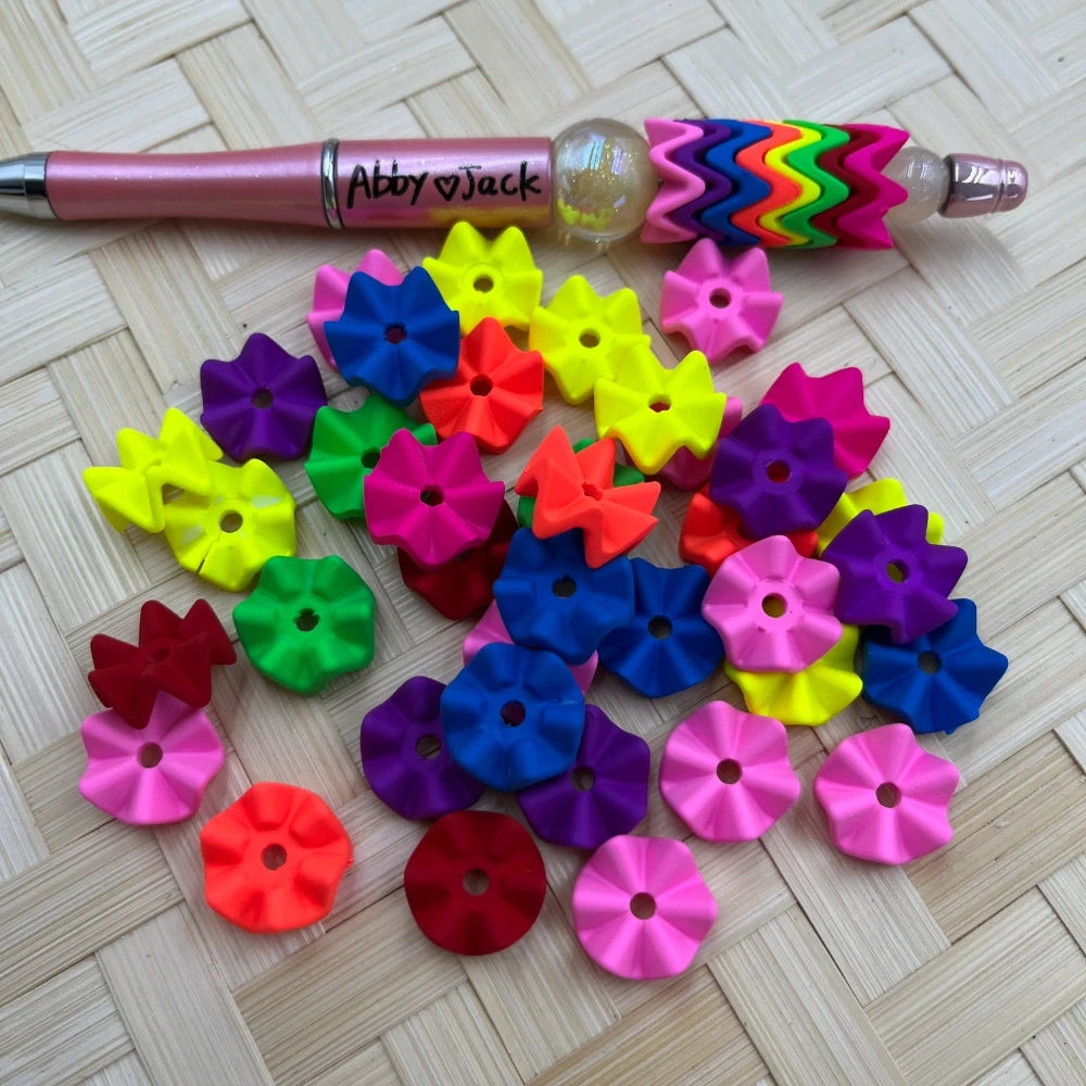 Assorted neon multicolor flower petal acrylic spacer beads in yellow, pink, blue, green, purple, red and orange scattered on a woven surface next to a beadable pen decorated with the same spacers.