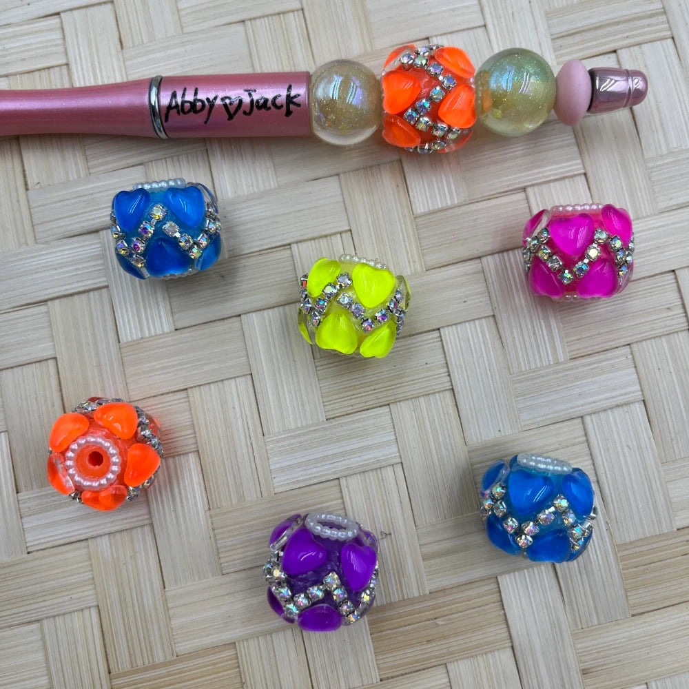Six colorful neon fancy beads decorated with heart-shaped cabochons and AB rhinestones in blue, yellow, orange, purple, pink, and teal, displayed on a woven mat beside an Abby Jack beadable pen.