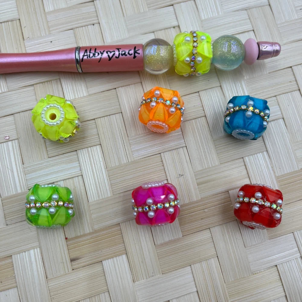 Colorful neon fancy chunky beads with rhinestone band and pearl accents in yellow-green, orange, teal, green, pink, and red, shown on a woven surface next to a beaded pen labeled Abby Jack.