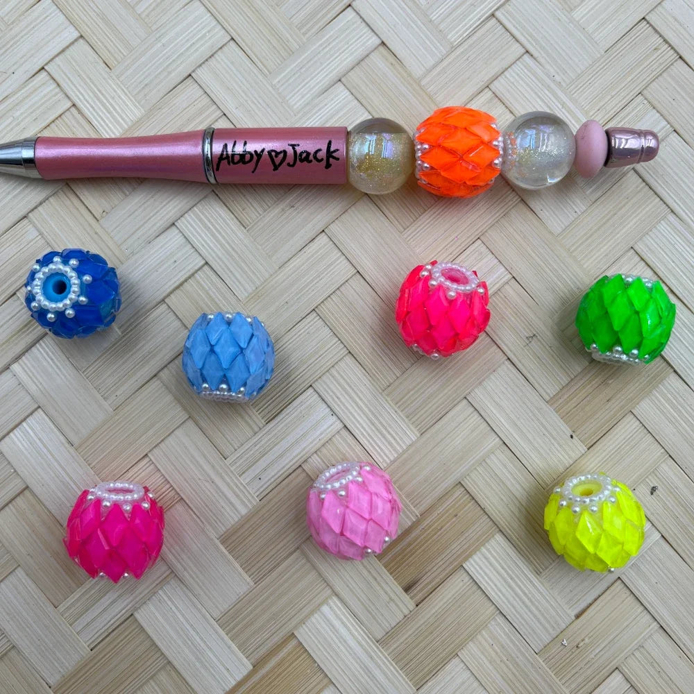 Assorted neon scallop-textured fancy beads with pearl rim detail in blue, light blue, hot pink, pink, neon green, yellow, and orange, displayed alongside a beadable pen on a woven bamboo surface.