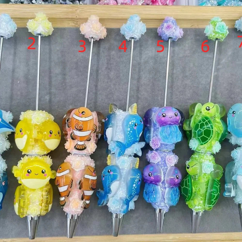 Decorative clay beadable pens with ocean animal characters including yellow pufferfish, clownfish, blue narwhal, purple jellyfish, green sea turtle, and blue dolphin, each topped with colorful flower embellishments on silver metal pens displayed on a wooden board.