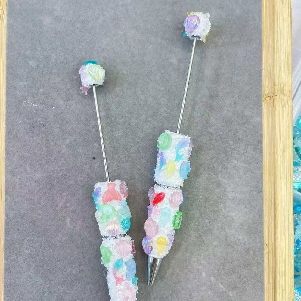 Two handmade clay beadable pens decorated with pastel ocean shell, mermaid tail, and starfish clay beads in pink, green, blue, and purple on a gray surface.