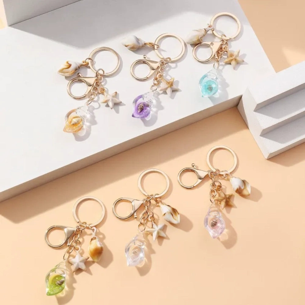 Six ocean-themed keychain charms featuring resin crystal shells in green, purple, blue, clear, and pink, with mini starfish and natural shell accents on gold-tone rings and lobster clasps.