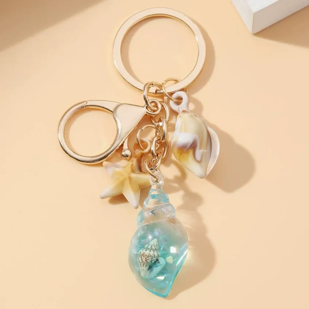 Six ocean-themed keychain charms featuring resin crystal shells in green, purple, blue, clear, and pink, with mini starfish and natural shell accents on gold-tone rings and lobster clasps. - 10