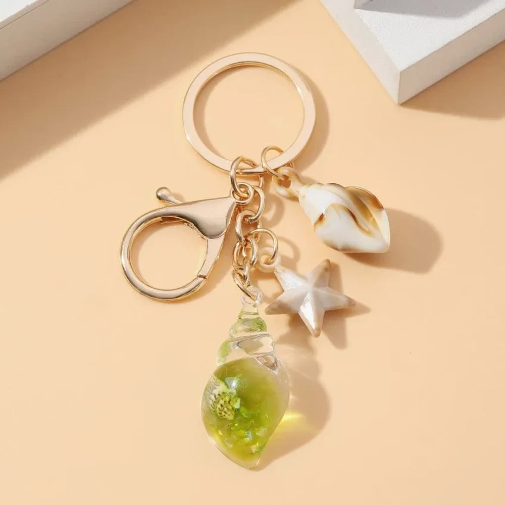 Six ocean-themed keychain charms featuring resin crystal shells in green, purple, blue, clear, and pink, with mini starfish and natural shell accents on gold-tone rings and lobster clasps. - 11